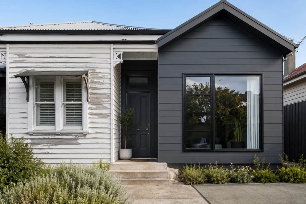 Front yard view showing an older white weatherboard house beside a dark grey composite cladding extension and concrete steps